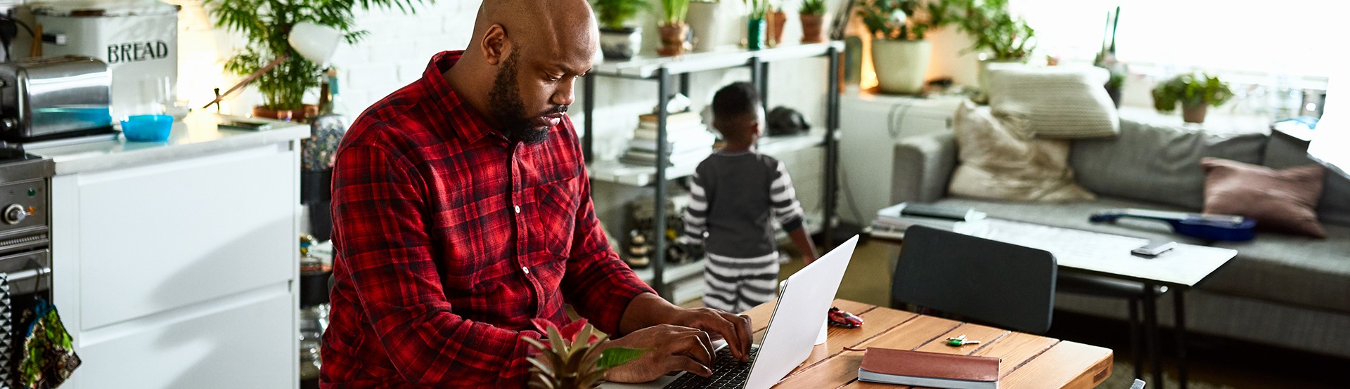 man working from home on a laptop surrounded by house plants with his son playing in the background